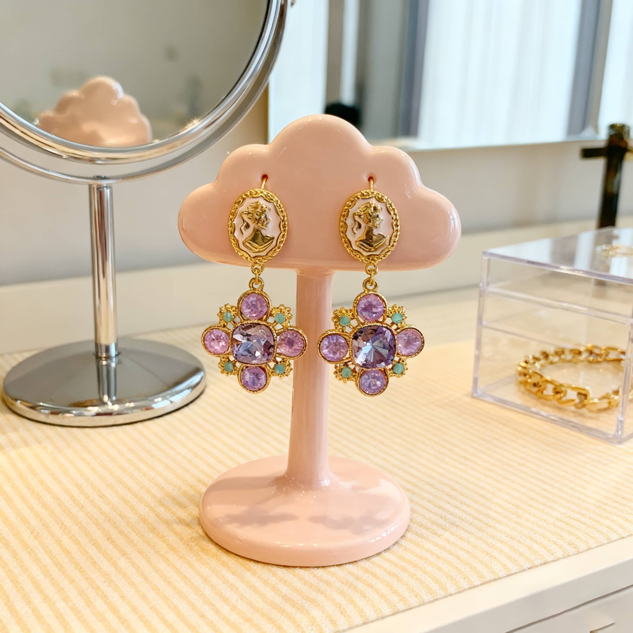Elegant baroque cameo floral earrings in gold with purple stones, displayed on a pink cloud-shaped stand