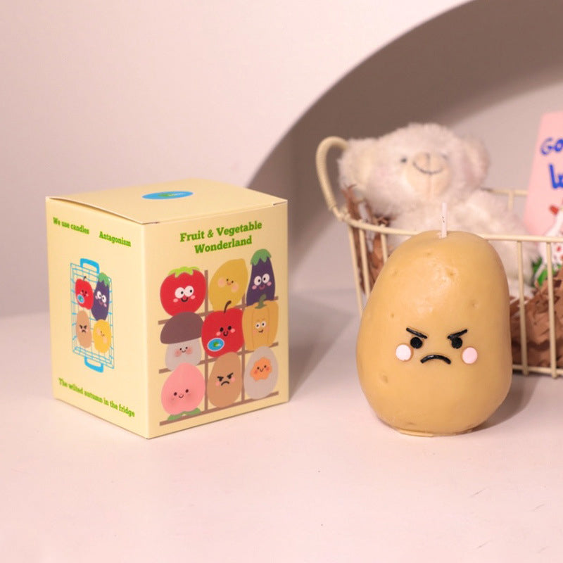 Hyper realistic food candle shaped like an angry potato with cartoon face next to colorful fruit and vegetable box