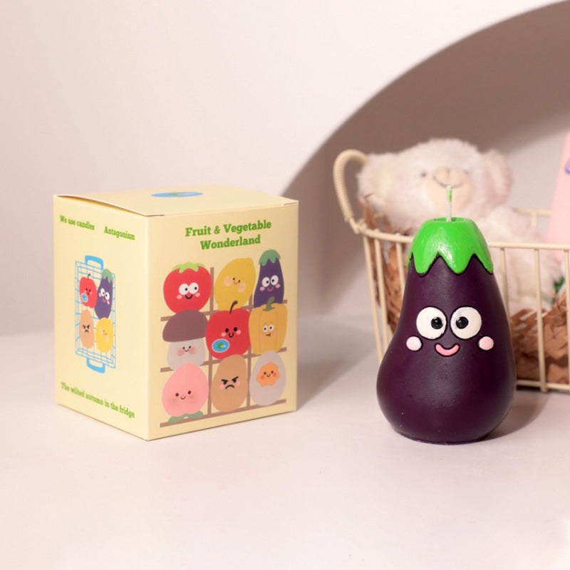 Hyper realistic food candle shaped like a smiling eggplant with green top next to colorful fruit and vegetable box