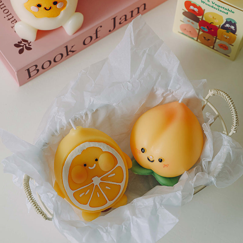 Two hyper realistic food candles shaped like a smiling peach and an orange slice in a white basket with tissue paper