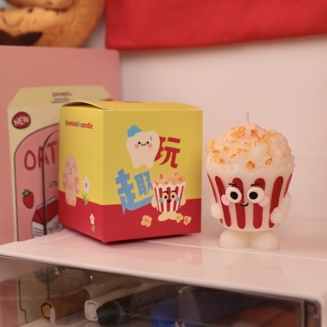 Hyper realistic food candle shaped like a popcorn bucket with cartoon eyes beside colorful packaging
