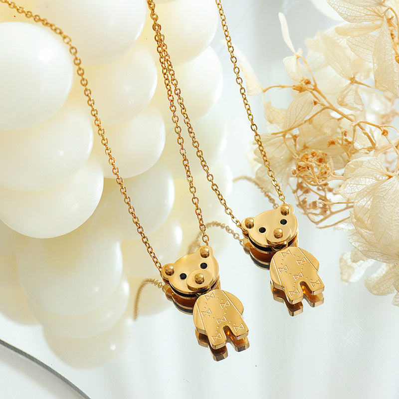 Two gold pendants on delicate chains featuring a golden teddy bear necklace on a reflective surface with dried flowers