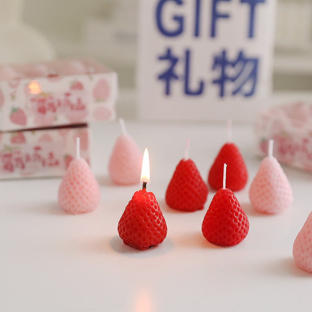 Red and pink self-love aromatherapy candle shaped like strawberries with one lit candle on white surface