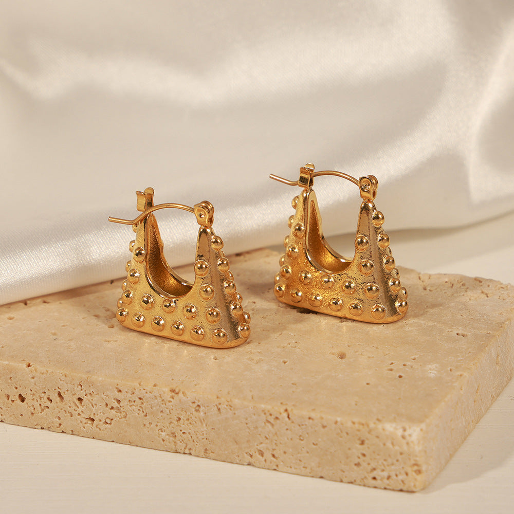 Geometric gold earrings with studded texture displayed on a stone platform with satin background