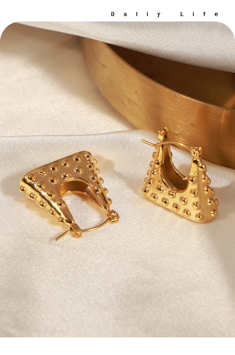 Geometric gold earrings with textured stud detailing on a soft fabric background